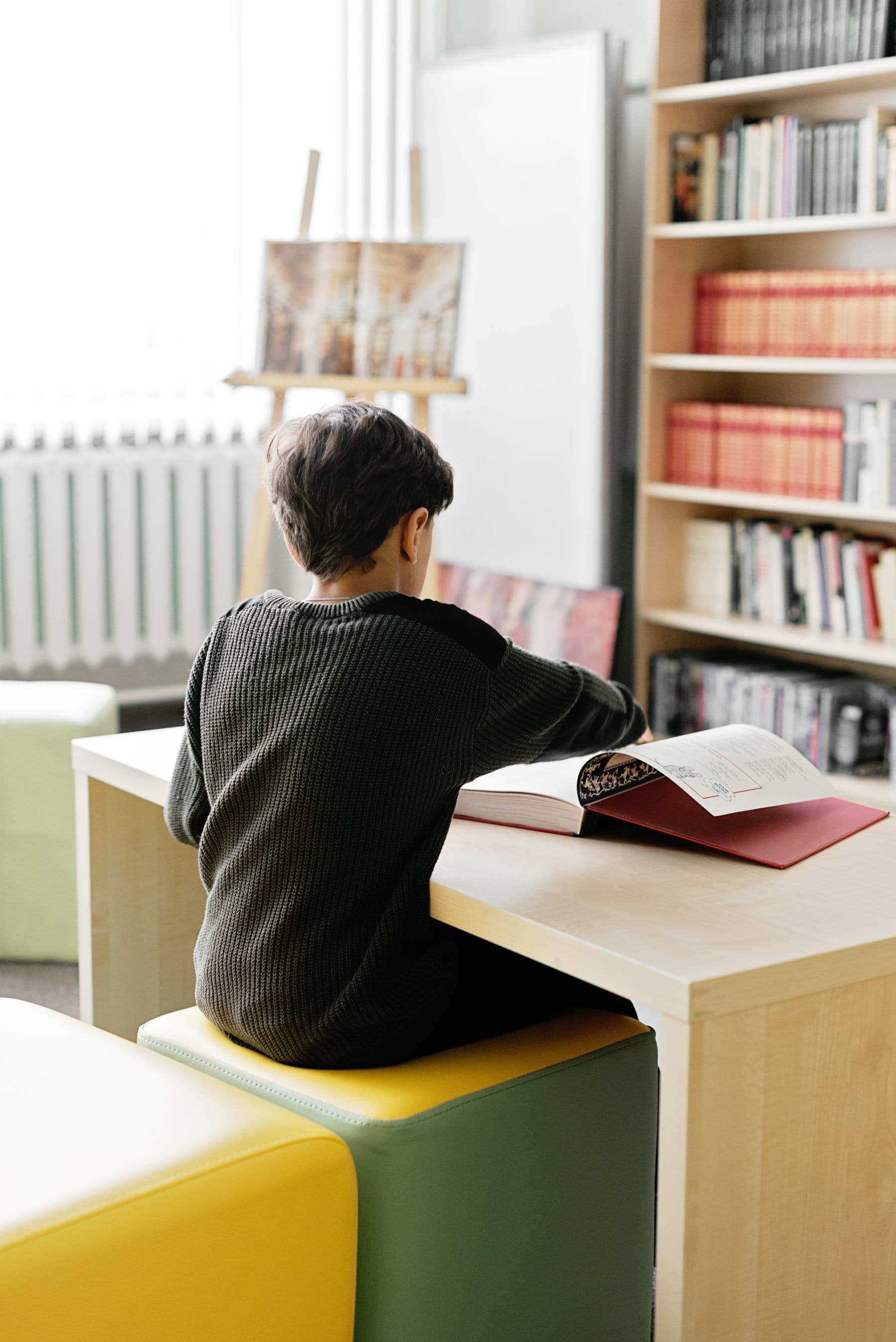 A child reading at a desk beside a bookshelf, seen from behind.
