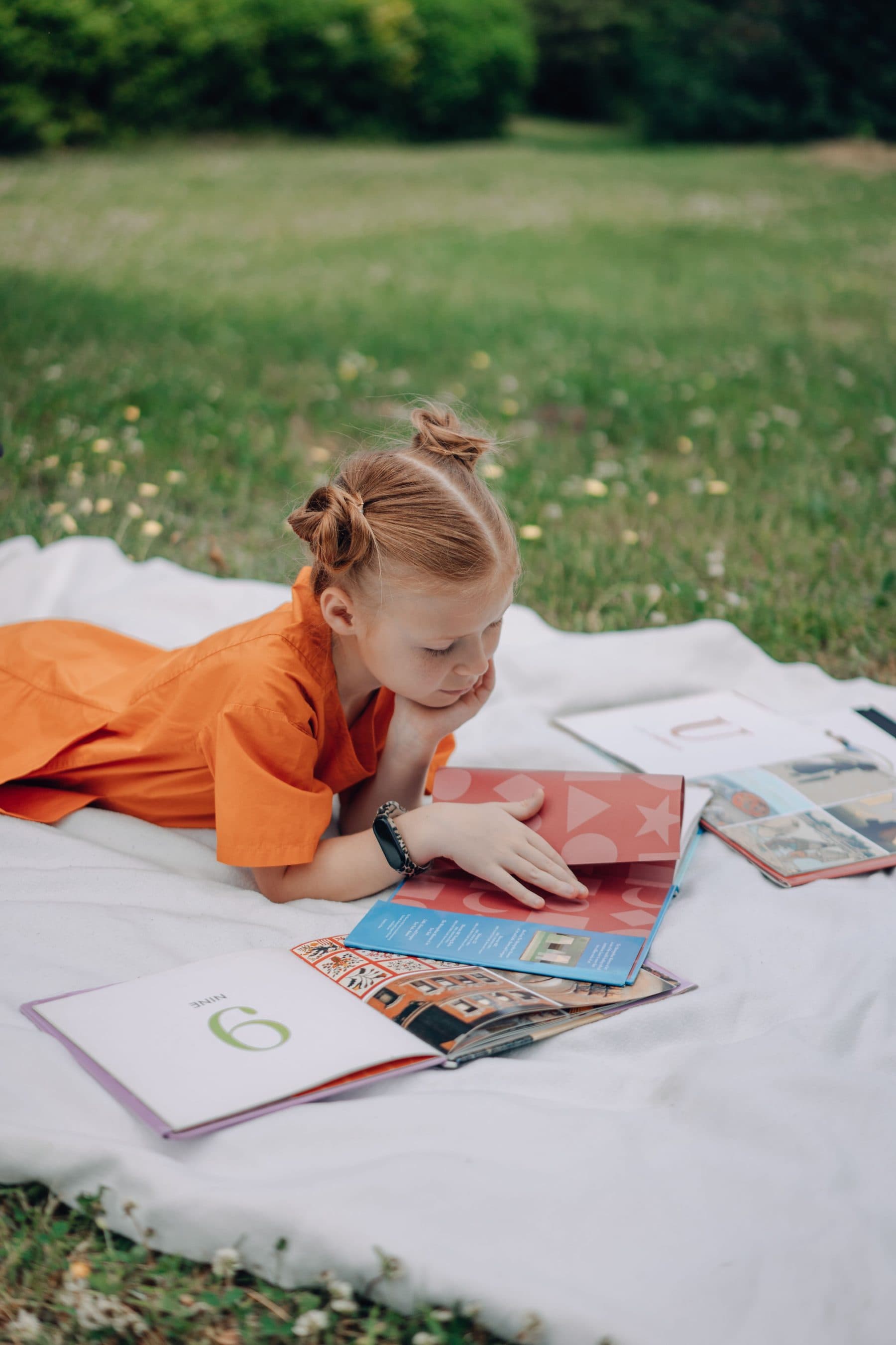 A child lying on a blanket in a meadow, chin in hand, absorbed in an open picture book with more books spread open nearby.