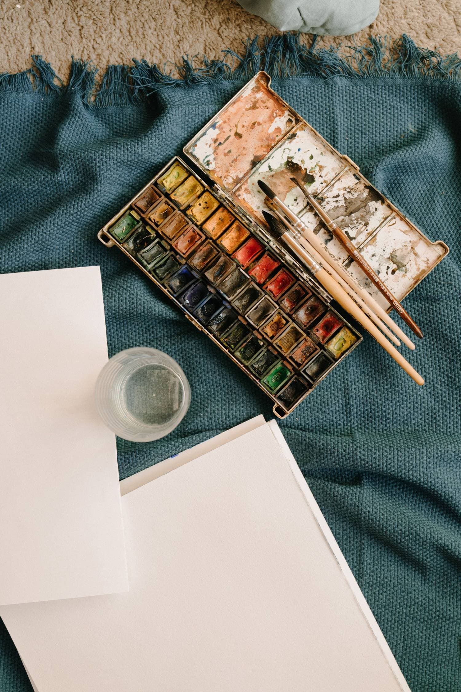 An open box of watercolors, brushes and a sheet of paper laid out on cloth.