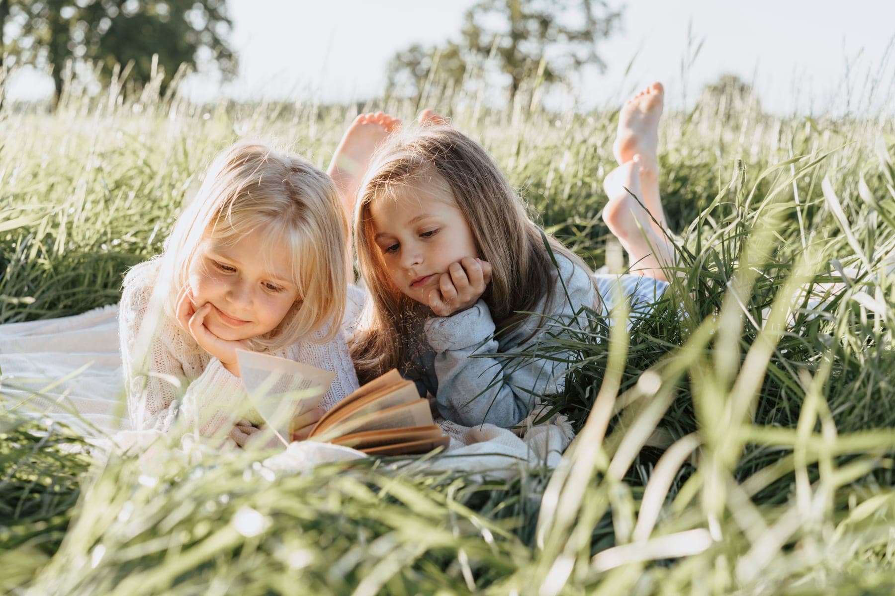 Two children lying on a blanket in long summer grass, reading a book together.